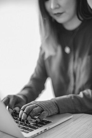 Una donna asiatica disabile scrive con il laptop e indossa i guanti di compressione. A fuoco sono le mani e la tastiera.