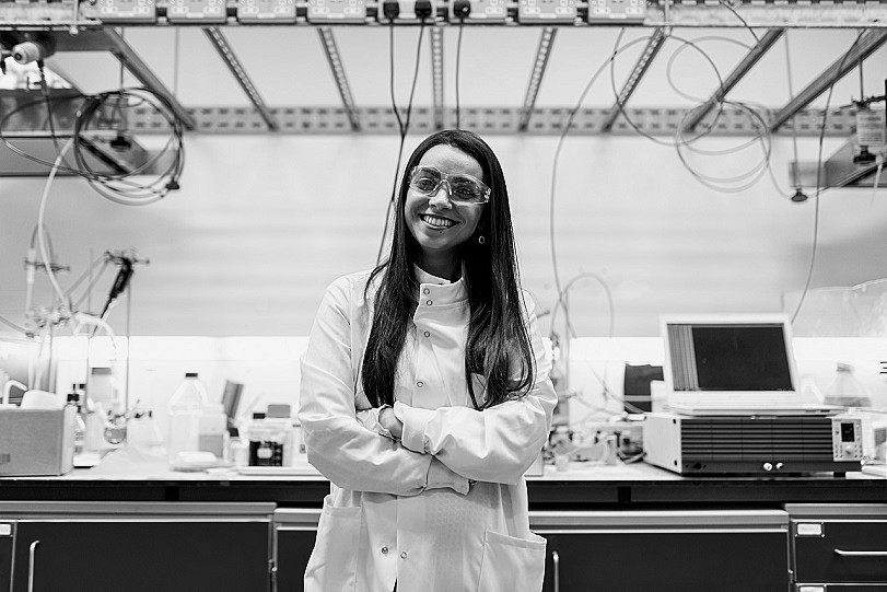 una donna con i capelli lunghi, il camice, gli occhiali protettivi sorride in un laboratorio di ingegneria chimica