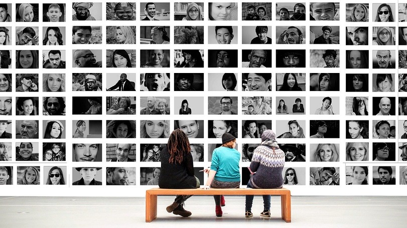 tre persone sono sedute su una panca in un museo e guardano un'opera composta da moltissime foto in bianco e nero di persone giovani, anziane, donne, uomini, nere, bianche, asiatiche. il linguaggio inclusivo rappresenta tutte le comunità meno rappresentate